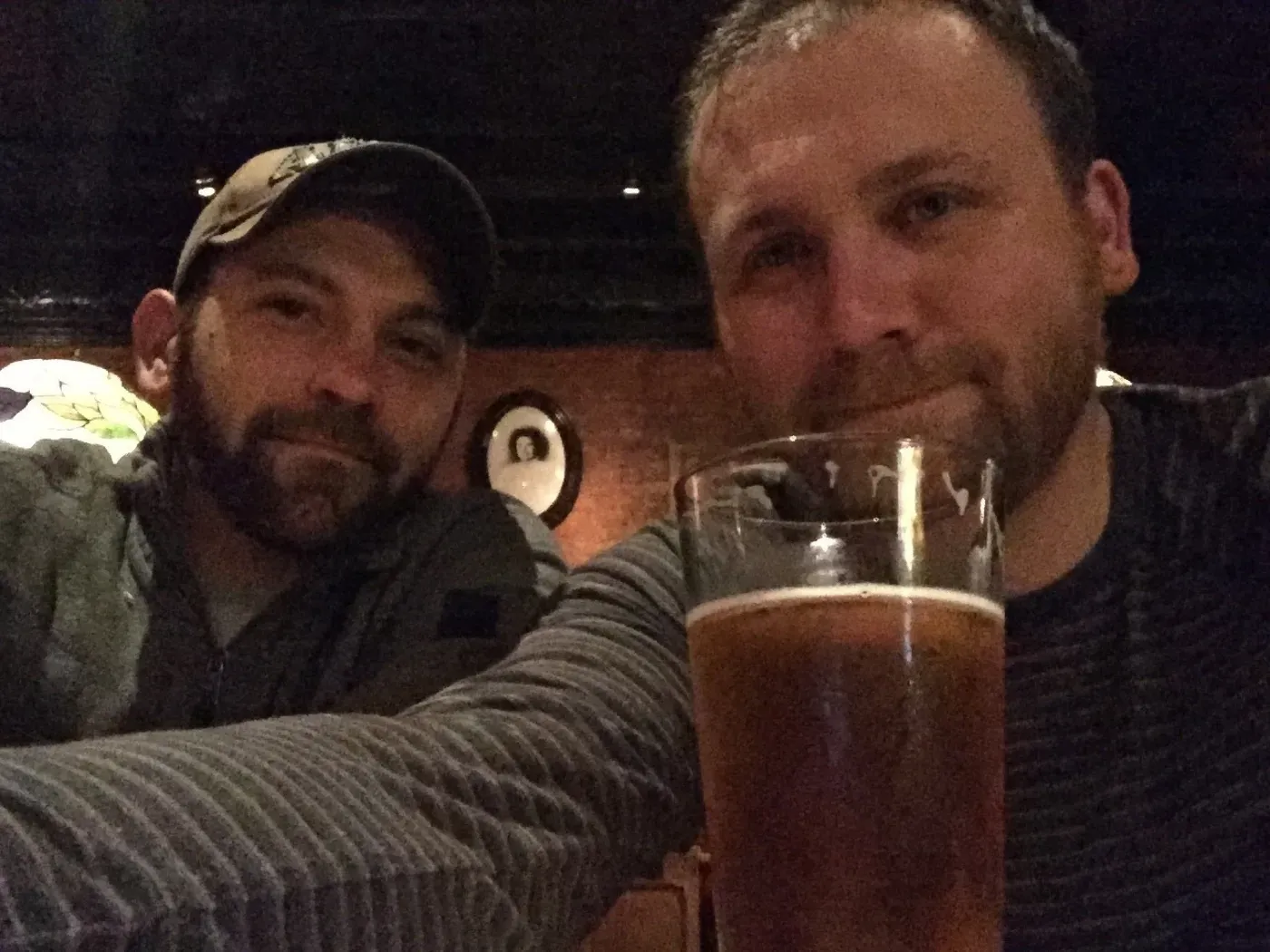 Two men are sitting together in a dimly lit setting, with one of them holding a glass of beer in the foreground.
