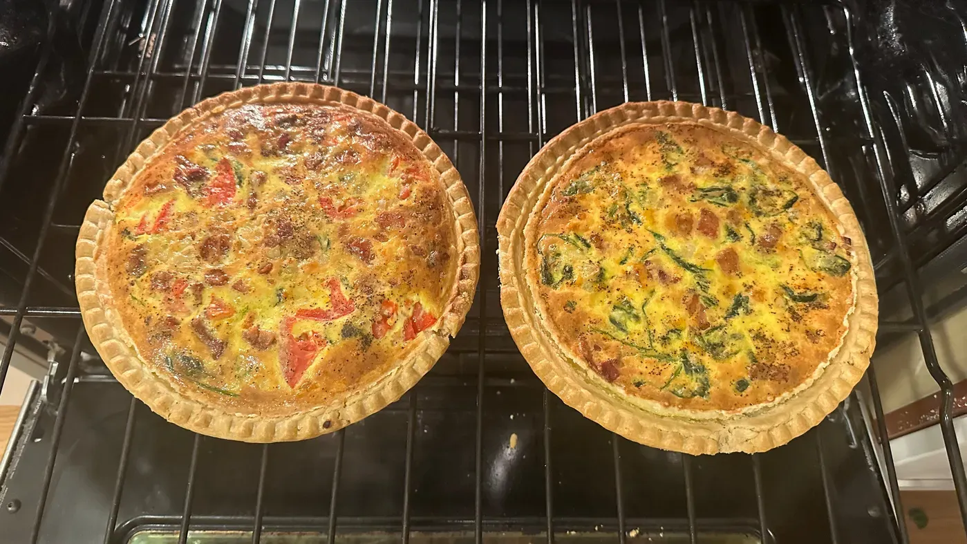 Two baked quiches with golden crusts and various fillings are on an oven rack.