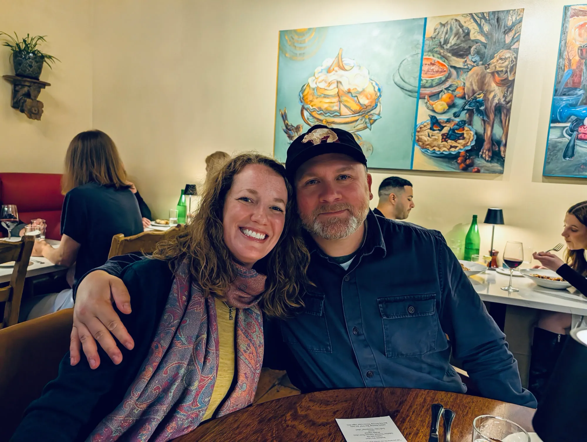 A couple doing a side hug at a restaurant table with other people at tables behind them and art on the wall.
